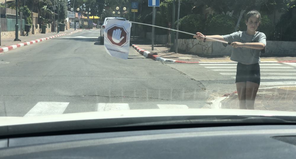 נערה עומדת בשפת מעבר החצייה ומושיטה את התמרורון אל הכביש, כפי שנראה מתוך רכב מתקרב.